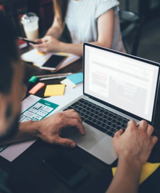 Bulanık ekin adam modern dizüstü bilgisayarda çalışıyor ve arka planda kağıtlar, malzemeler ve iş arkadaşıyla masada otururken daktilo yazıyor.