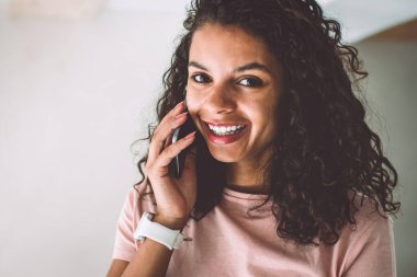 Kıvırcık saçlı, gülümseyen ve telefona bakan neşeli, esmer tenli hippi kızın yarım boy portresi teknoloji iletişimi ve insan konsepti. Akıllı telefondan arayan mutlu kadın.