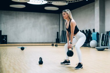 Son moda spor kıyafetli genç bir kadının Kettlebell 'le spor yapıp aynaya bakarken spor salonunda çömelmesi.