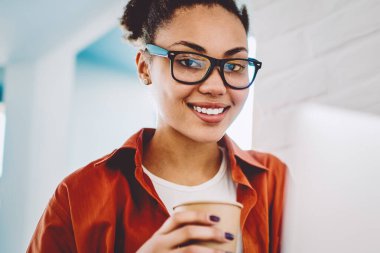 Optik gözlüklü, serbest çalışan, elinde yemek fincanı tutan ve reklam alanında kahve molası veren yetenekli bir kadın portresi. Kafeteryada dizüstü bilgisayarlı mutlu koyu tenli hippi kız.