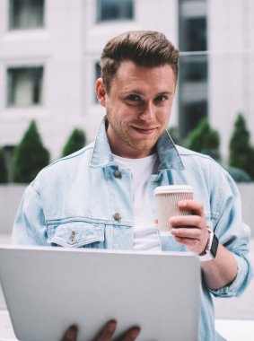 Kot ceketli genç beyaz adam dizüstü bilgisayar tutuyor ve sokakta durup kameraya gülümserken bir fincan kahve içiyor.