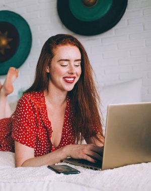 Romantik, mutlu, kırmızı saçlı, parlak noktalı elbiseli bir kadın evde akıllı telefon ve dizüstü bilgisayar kullanırken rahat bir yatakta uzanıyor.