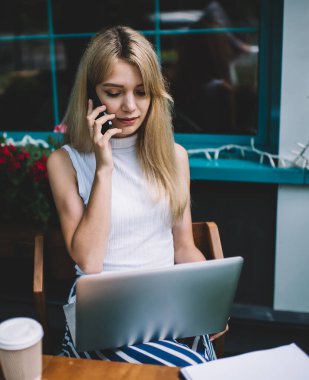 Sarışın, ciddi, gülümseyen, kolsuz tişörtlü, cep telefonuyla konuşan, şehirdeki sokak kafeteryasında dizüstü bilgisayarla oturan zeki bir kadın.