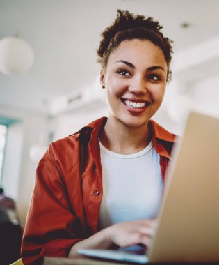 Neşeli hippi kız modern netbook ile birlikte çalışma alanında oturuyor ve komik mesajlar alıyor, olumlu siyah kadın kafasını çeviriyor ve kafenin içinde öğrenme sırasında gülümsüyor.