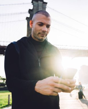 Siyah spor giyim giymiş, keyifli atletik genç adam şehir köprüsünde arka plandaki metal çitlerde akıllı telefonuna dokunuyor.