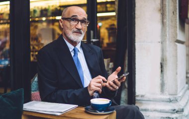 Gözlüklü, takım elbiseli, akıllı telefonlu yaşlı bir adam cama yaslanmış ve kahve fincanıyla yan yana oturuyor.