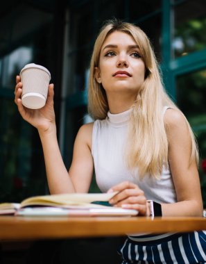 Genç, uzun saçlı, elinde kahve fincanı tutan ve kahve dükkanının önündeki verandada oturup gözlerini başka yöne çeviren bir kadın.