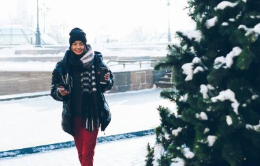 Kağıt bardak ve telefonla kışın gülümseyen genç bayan karlı caddede Noel ağaçlarının yanında yürüyor ve kameraya bakıyor.
