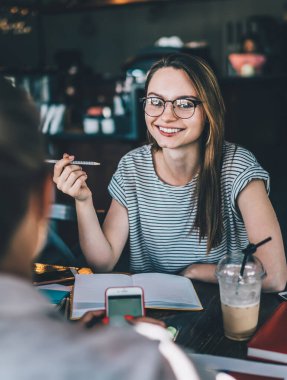 Gözlüklü hoş, sıradan bir kadın kafeteryada oturup içerken kız arkadaşıyla konuşuyor ve not defterlerini inceliyor.