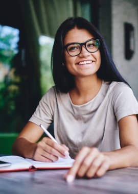 Gözlüklü neşeli esmer kadının portresi. Kameraya bakıyor, not defterinde planlar yapıyor. Kafenin içinde oturuyor, güzel ve yaratıcı fikirler yazan bir kadın.