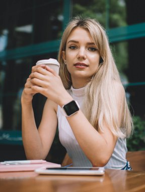 Beyaz kolsuz balıkçı yaka giyen, modern kol saati takan, kahve dükkanında elinde kahve fincanıyla oturan ve kameraya bakan sarışın genç bir kadın.