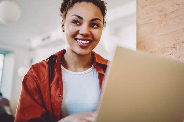 Neşeli hippi kız modern netbook ile birlikte çalışma alanında oturuyor ve komik mesajlar alıyor, olumlu siyah kadın kafasını çeviriyor ve kafenin içinde öğrenme sırasında gülümsüyor.