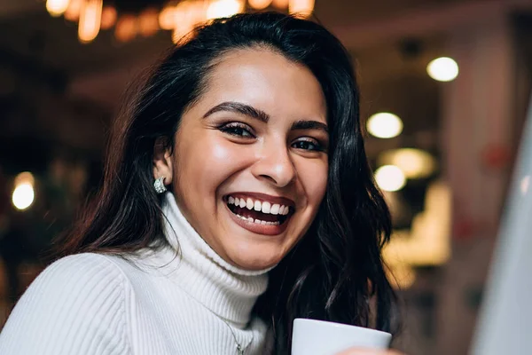 Cheerful white toothed smart lady with dark long hair in white jumper sitting at table with laptop holding cup of coffee in modern coffee shop