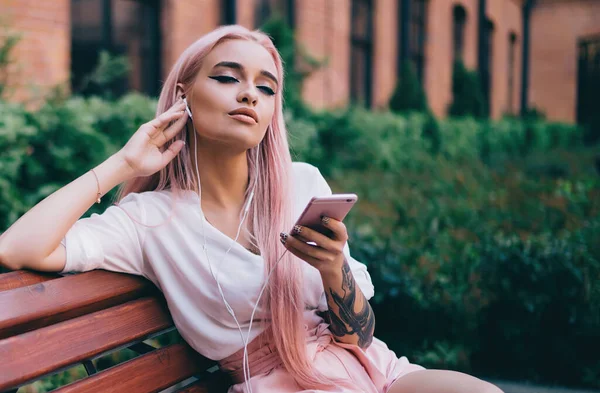 Young lady in white shirt and pink shorts with long pink hair listening to music on smartphone sitting on wooden bench in street opposite of brick building