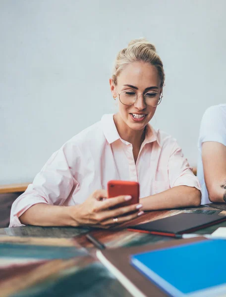 Tattooed and bearded brunet guy and blond young woman sitting at table with laptop and notepads sharing smartphone and having fun