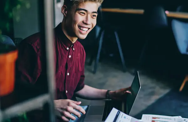 Positive chinese hipster student laughing while holding modern laptop in hand.Cheerful asian young man dressed in stylish casual wear smiling during remote work at digital netbook and paper reports