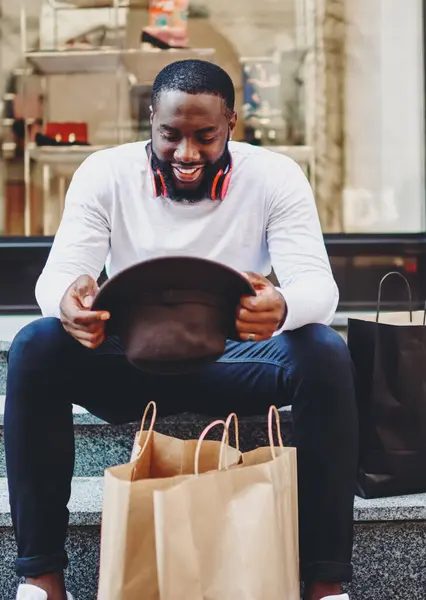 Front view of cheerful dark skinned male shopaholic resting on stairs after shopping time looking on fashionable hat and feeling good from bought things, happy hipster guy with paper bags outdoors
