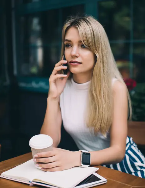 Young blond woman wearing sleeveless turtleneck sitting at table outside coffee shop and talking on phone while looking away with cup of coffee in hand