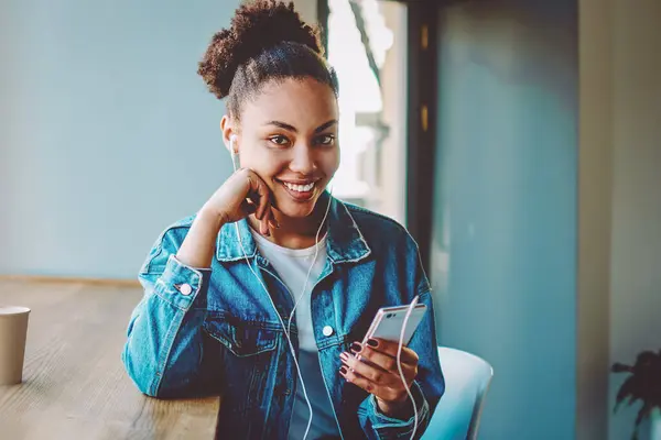 Portrait of excited smiling hipster girl dressed in stylish apparel looking at camera while listening favourite song from motivation playlist on cellular phone, happy woman enjoying audio book
