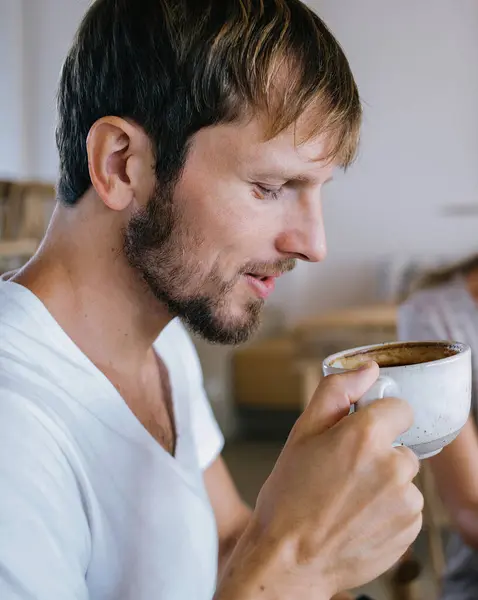 Güzel milenyum erkeğine odaklanın. Sıcak, keskin kahvenin tadını çıkarırken arka planda bulanık bir kadın kağıda yazı yazıyor ve gülümsüyor.