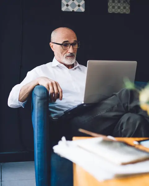Senior bearded bald man in white shirt and black pants with glasses working on laptop while sitting in living room