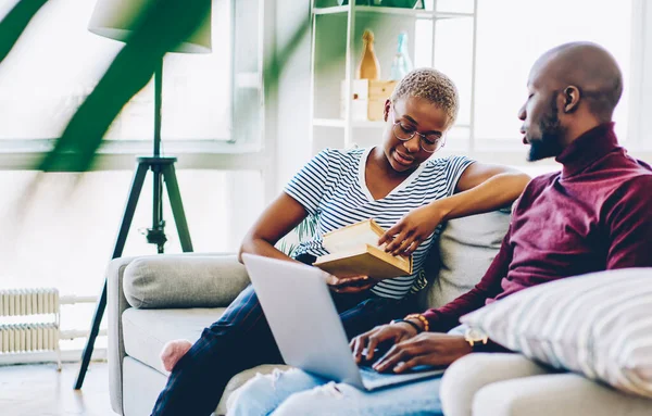 Sevgili Afrikalı Amerikalı çift, kız arkadaşı şık bir şekilde oturma odasında oturmuş kitap okurken dizüstü bilgisayarını kullanan ve evde dinlenerek vakit geçiriyorlar.
