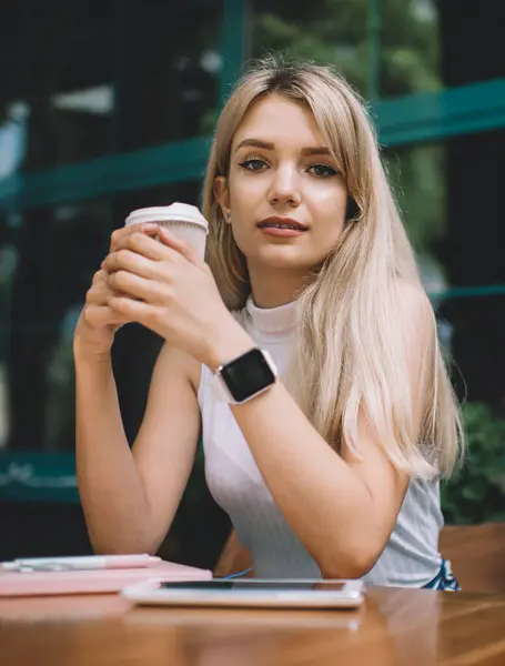 Beyaz kolsuz balıkçı yaka giyen, modern kol saati takan, kahve dükkanında elinde kahve fincanıyla oturan ve kameraya bakan sarışın genç bir kadın.