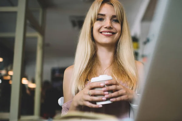 Low angle of young woman with long blond hair wearing sleeveless clothing while sitting in coffee shop and holding cup of coffee and smiling at camera