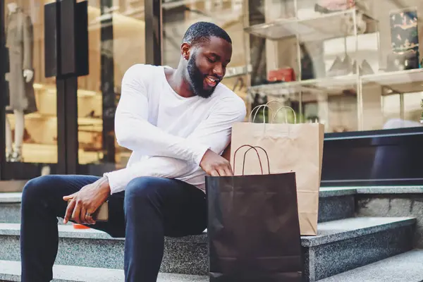 Smiling dark skinned hipster guy dressed in long sleeve shirt looking at shopping bags checking purchases, positive african american male happy about buying clothing in wear store with good discount