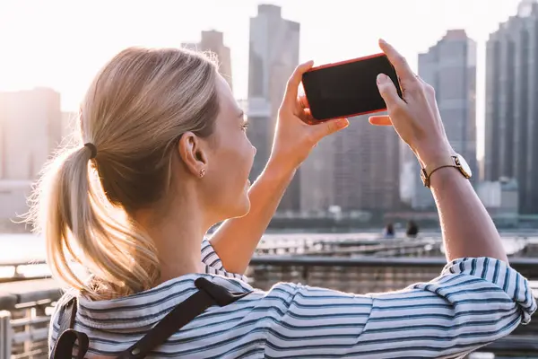 Back view of millennial woman in casual wear shooting video of New York landscape using smartphone main camera for clicking pictures, female tourist testing cellphone while taking photos of city