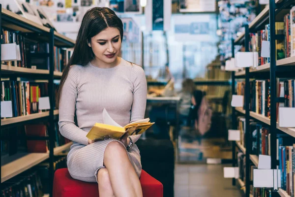 Bej elbiseli güzel bir kadın puf üzerinde oturuyor ve üniversite kütüphanesinde kitaplıkların yanında otururken sarı kapaklı kitap okuyor.