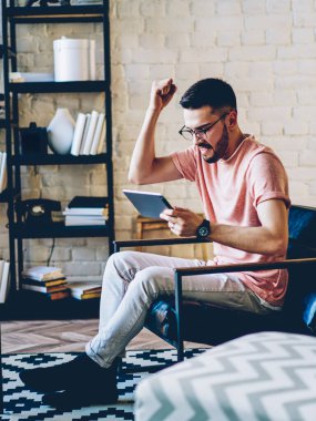 Modern dairede rahat bir sandalyede oturan genç hippi modern dijital tabletle online oyun oynarken zaferi kutluyor ve inanılmaz haberler okuyor.