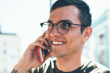 Gözlüklü ve dövmeli gülümseyen adam güneşli bir günde şehirde yürüyen arkadaşıyla sohbet etmek için telefonunu kullanıyor.