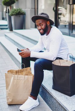 Ağ mağazası servisinden geribildirim bekleyen Afro-Amerikalı bir adamın portresi merdivenlerde oturmuş akıllı telefondan alışveriş sonrası dinlenen şık şapkalı, pozitif esmer bir adam.