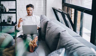 Modern tasarlanmış oturma odasında kanepeye uzanan neşeli hippi dizüstü bilgisayar ve kablosuz internetten alışveriş yapıyor elinde kredi kartı tutan adam evdeki web mağazasında dinlenme yerinin parasını ödüyor.