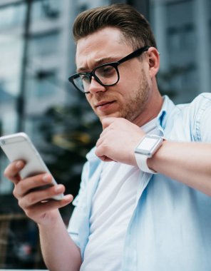 Akıllı saat ve gözlüklü yakışıklı milenyum erkeğinin sokak terasında otururken konsantrasyonlu akıllı telefon kullanmasından.