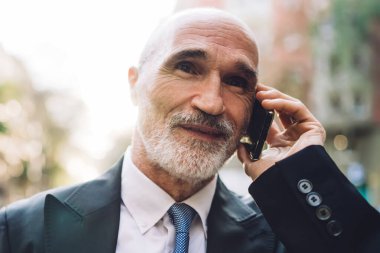 Gülümseyen, takım elbiseli, beyaz sakallı, sokakta yürürken telefonla konuşan, bulanık binalara bakan yaşlı, yakışıklı bir erkek.