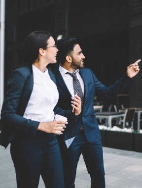 Katı siyah takım elbiseli çok ırklı iş arkadaşları yaz aylarında şehir merkezinde bir apartmanı işaret ederek birbirleriyle konuşuyorlar.