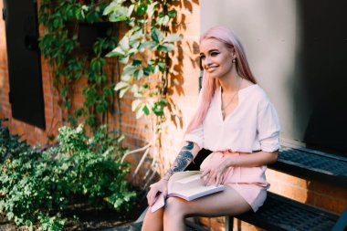 Pembe saçlı ve dövmeli şık yaratıcı bir kadın. Elinde kitapla tuğladan duvarlar, siyah kapılar ve yeşil bitkilerle oturuyor.