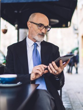 Takım elbiseli, sakallı, kahve fincanı, gazete ve sokak tabletinde mesajlaşan kıdemli bir adam.