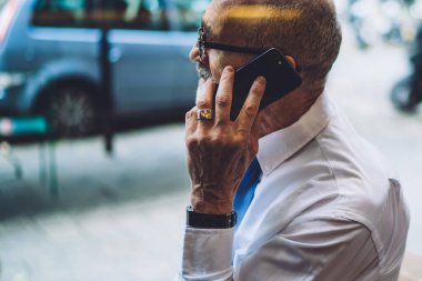 Beyaz tişörtlü, kısa saçlı yaşlı işadamının yan görüntüsü şehir sokaklarının ve yolların arka planında oturup telefonla konuşması.
