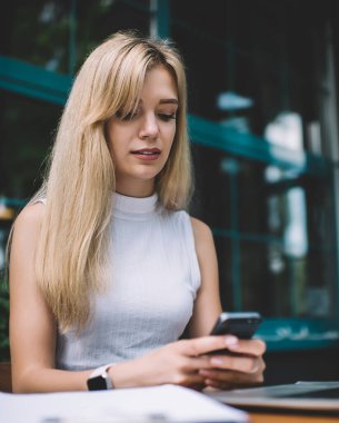 Kolsuz balıkçı yaka kazak giymiş, elinde akıllı bir telefon tutan ve gündüz kahve dükkanının önünde dizüstü bilgisayarla oturan genç sarışın bir kadın.