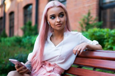 Beyaz tişörtlü, pembe şortlu, uzun pembe saçlı, cep telefonuyla müzik dinleyen caddedeki ahşap bankta oturan sıska kadın.