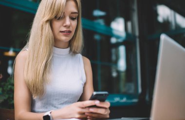 Kolsuz balıkçı yaka kazak giymiş, elinde akıllı bir telefon tutan ve gündüz kahve dükkanının önünde dizüstü bilgisayarla oturan genç sarışın bir kadın.