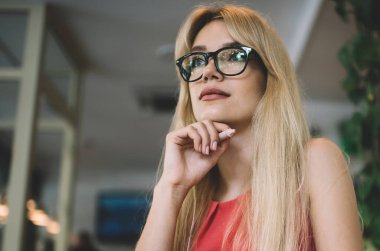 Gözlüklü zeki genç bir kadın ve bir bardak içecekle kafeterya masasında otururken gözlerini kaçırıp düşünecek.