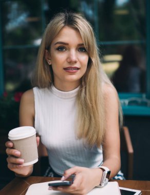 Bir fincan kahve içerken ve kahve masasında otururken cep telefonu kullanan uzun saçlı genç bir kadın.