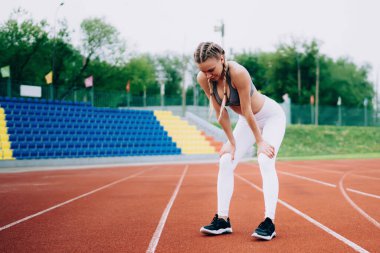 Sportif olarak zayıf yetişkin bir kadın, piste çıkıp dizlerinin üstüne çöker ve stadyum koltuklarına bakarak egzersiz yaptıktan sonra nefes almaya çalışır.