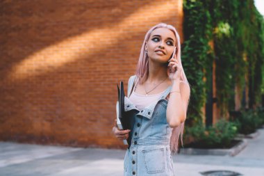 Gülümseyen ve arkasını kollayan defterleri olan neşeli genç bir kadın kaldırımda dikilip telefonlarına cevap veriyor.