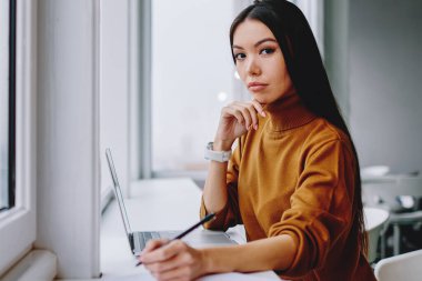 Ciddi esmer kadın çalışanın portresi dizüstü bilgisayarla oturmuş araştırma ve muhasebe yapıyor, çekici kendine güvenen iş kadını kameraya bakıyor ve pencerenin yanında notlar alıyor ve planlar yapıyor.