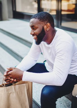 Gülümseyen afro-amerikan erkek müşteriler mağazanın merdivenlerinde çantalarla oturuyorlar, neşeli esmer tenli hippi adam alışveriş yaptıktan ve alışveriş yaptıktan sonra sokak tanıtım alanında eğleniyor.